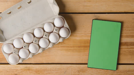 Overhead-Shot-Of-Twelve-Eggs-In-Cardboard-Box-Being-Opened-On-Wooden-Table-With-Digital-Tablet