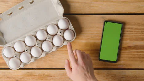 Fotografía-Cenital-De-Una-Persona-Que-Elige-Un-Huevo-De-Una-Caja-De-Cartón-En-Una-Mesa-De-Madera-Con-Teléfono-Móvil