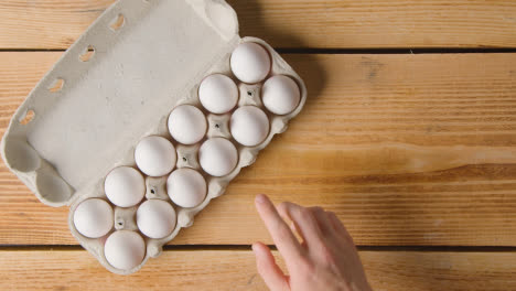 Overhead-Shot-Of-Person-Choosing-From-Box-Of-Twelve-Eggs-Open-On-Wooden-Table