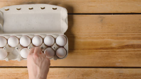 Overhead-Shot-Of-Person-Choosing-From-Box-Of-Twelve-Eggs-Open-On-Wooden-Table