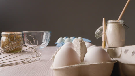 Foto-De-Estudio-De-Ingredientes-Y-Utensilios-Para-Hornear-En-La-Encimera-De-La-Cocina-Con-Una-Persona-Recogiendo-Huevos