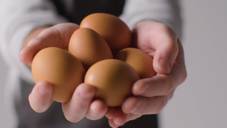 Close-Up-Studio-Shot-Of-Person-In-Apron-Holding-Up-Handful-Of-Brown-Eggs-To-Camera