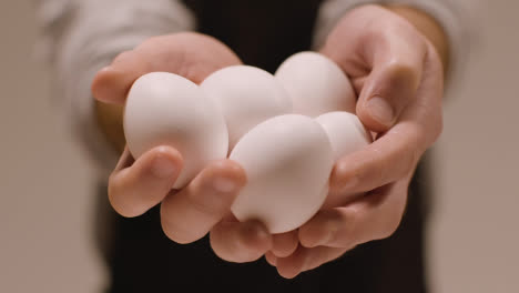 Cerrar-Foto-De-Estudio-De-Persona-En-Delantal-Sosteniendo-Un-Puñado-De-Huevos-Blancos-A-La-Cámara