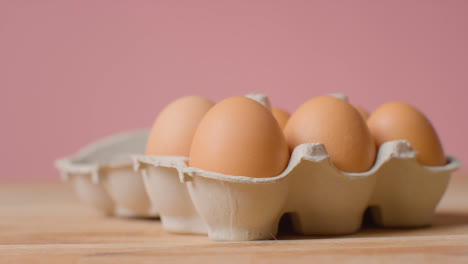 Studio-Shot-Of-Person-Opening-Cardboard-Box-Containing-Brown-Eggs-Against-Pink-Background