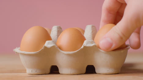 Studio-Shot-Of-Person-Choosing-From-Open-Cardboard-Box-Containing-Brown-Eggs-Against-Pink-Background