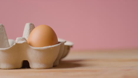 Studio-Shot-Of-Person-Taking-Brown-Egg-From-Open-Cardboard-Box-Against-Pink-Background
