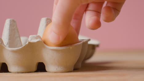 Studio-Shot-Of-Person-Putting-Brown-Egg-Into-Open-Cardboard-Box-Against-Pink-Background