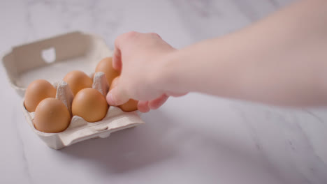 Person-Choosing-Egg-From-Open-Cardboard-Box-On-Marble-Work-Surface-Background
