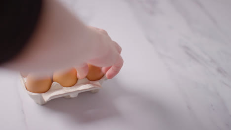 Person-Choosing-Egg-From-Open-Cardboard-Box-On-Marble-Work-Surface-Background