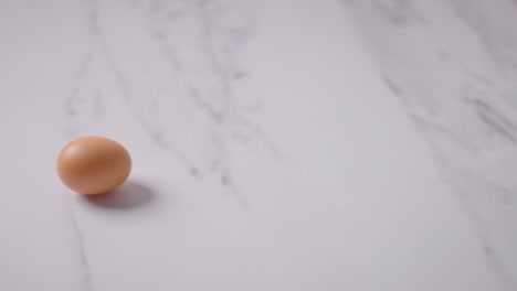 Studio-Shot-Of-Hand-Picking-Up-Single-Brown-Egg-On-Marble-Work-Surface-Background
