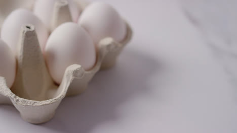 Studio-Shot-Of-Person-Putting-White-Egg-Into-Open-Cardboard-Box-On-Marble-Work-Surface-Background