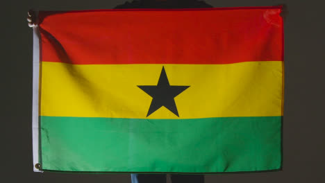 Foto-De-Estudio-De-Una-Persona-Anónima-O-Un-Aficionado-A-Los-Deportes-Que-Sostiene-La-Bandera-De-Ghana-Contra-Un-Fondo-Negro