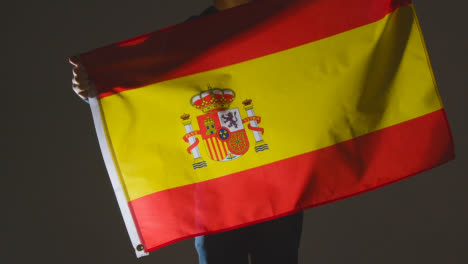 Studioaufnahme-Einer-Anonymen-Person-Oder-Eines-Sportfans,-Die-Die-Flagge-Spaniens-Vor-Schwarzem-Hintergrund-Schwenken