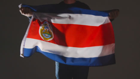 Studioaufnahme-Einer-Anonymen-Person-Oder-Eines-Sportfans,-Die-Die-Flagge-Von-Costa-Rica-Vor-Schwarzem-Hintergrund-Schwenken