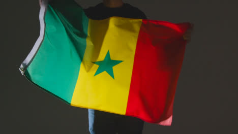Studio-Shot-Of-Anonymous-Person-Or-Sports-Fan-Waving-Flag-Of-Senegal-Against-Black-Background