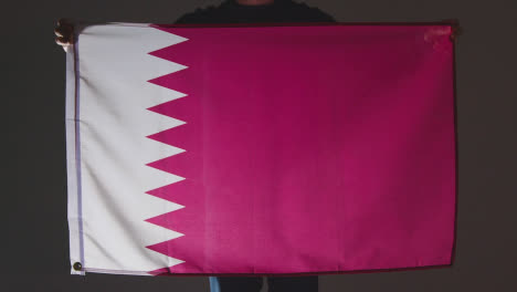 Studio-Shot-Of-Anonymous-Person-Or-Sports-Fan-Holding-Flag-Of-Qatar-Against-Black-Background