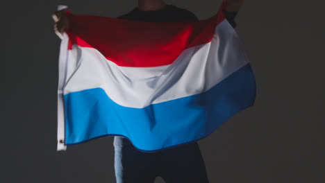 Studio-Shot-Of-Anonymous-Person-Or-Sports-Fan-Waving-Flag-Of-Netherlands-Against-Black-Background