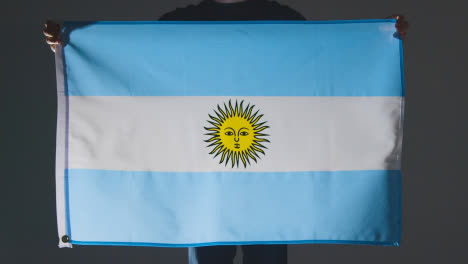Studio-Shot-Of-Anonymous-Person-Or-Sports-Fan-Holding-Flag-Of-Argentina-Against-Black-Background