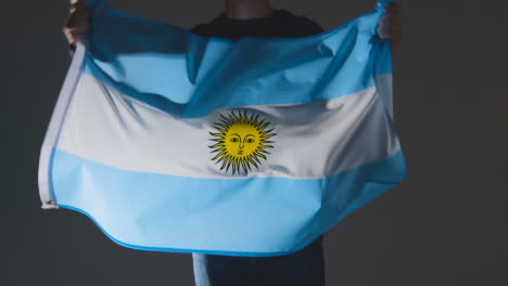 Studio-Shot-Of-Anonymous-Person-Or-Sports-Fan-Waving-Flag-Of-Argentina-Against-Black-Background
