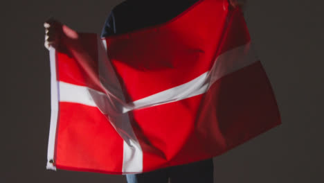 Studio-Shot-Of-Anonymous-Person-Or-Sports-Fan-Waving-Flag-Of-Denmark-Against-Black-Background