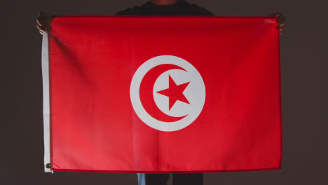 Studio-Shot-Of-Anonymous-Person-Or-Sports-Fan-Holding-Flag-Of-Tunisia-Against-Black-Background