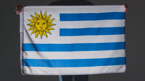 Foto-De-Estudio-De-Una-Persona-Anónima-O-Fanático-De-Los-Deportes-Que-Sostiene-La-Bandera-De-Uruguay-Contra-Fondo-Negro