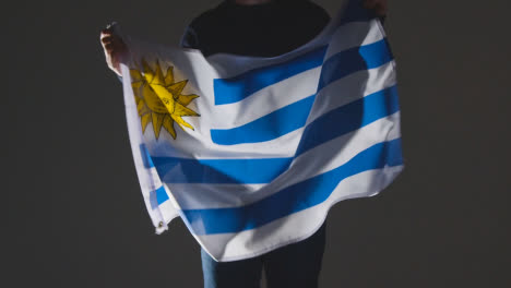 Studioaufnahme-Einer-Anonymen-Person-Oder-Eines-Sportfans,-Die-Die-Flagge-Uruguays-Vor-Schwarzem-Hintergrund-Schwenken