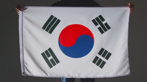 Foto-De-Estudio-De-Una-Persona-Anónima-O-Fanático-De-Los-Deportes-Que-Sostiene-La-Bandera-De-Corea-Del-Sur-Contra-Un-Fondo-Negro