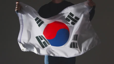 Foto-De-Estudio-De-Una-Persona-Anónima-O-Fanático-De-Los-Deportes-Ondeando-La-Bandera-De-Corea-Del-Sur-Contra-Fondo-Negro