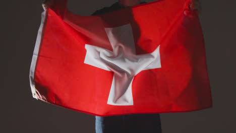 Studioaufnahme-Einer-Anonymen-Person-Oder-Eines-Sportfans,-Der-Die-Flagge-Der-Schweiz-Vor-Schwarzem-Hintergrund-Schwenkt