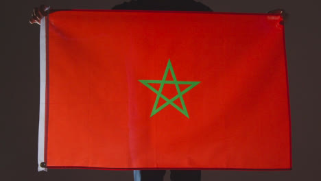 Foto-De-Estudio-De-Una-Persona-Anónima-O-Fanático-De-Los-Deportes-Que-Sostiene-La-Bandera-De-Marruecos-Contra-Fondo-Negro