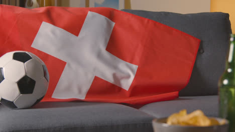 Die-Schweizer-Flagge-Hängt-Zu-Hause-über-Dem-Sofa,-Während-Der-Fussball-Im-Tv-1-Spielbereit-Ist