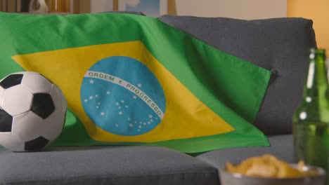 Flagge-Von-Brasilien-über-Dem-Sofa-Zu-Hause-Mit-Fußball-Bereit-Für-Das-Spiel-Im-Tv-1