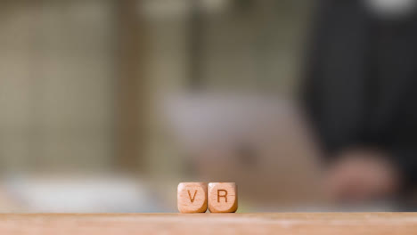 Business-Concept-Wooden-Letter-Cubes-Or-Dice-Spelling-VR-With-Office-Person-Working-At-Computer-In-Background