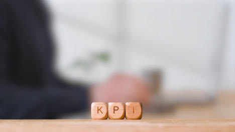Business-Concept-Wooden-Letter-Cubes-Or-Dice-Spelling-KPI-With-Office-Person-Working-At-Laptop-In-Background
