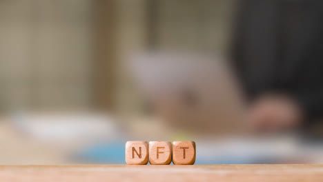 Business-Concept-Wooden-Letter-Cubes-Or-Dice-Spelling-NFT-With-Person-Working-On-Laptop-In-Office-In-Background