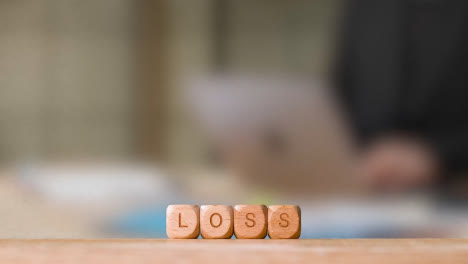 Business-Concept-Wooden-Letter-Cubes-Or-Dice-Spelling-Loss-With-Office-Person-Working-At-Laptop-In-Background
