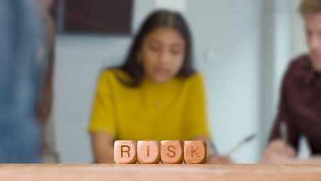 Business-Concept-Wooden-Letter-Cubes-Or-Dice-Spelling-Risk-With-Office-Meeting-In-Background