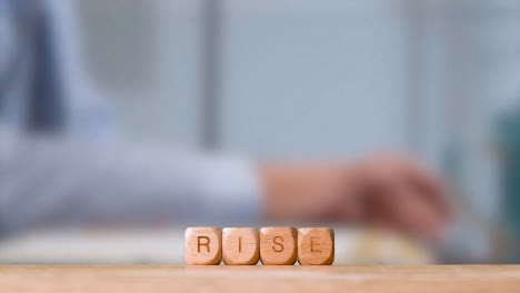 Business-Concept-Wooden-Letter-Cubes-Or-Dice-Spelling-Rise-With-Office-Person-Working-At-Laptop-In-Background