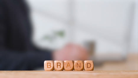 Business-Concept-Wooden-Letter-Cubes-Or-Dice-Spelling-Brand-With-Office-Person-Using-Mobile-Phone-In-Background