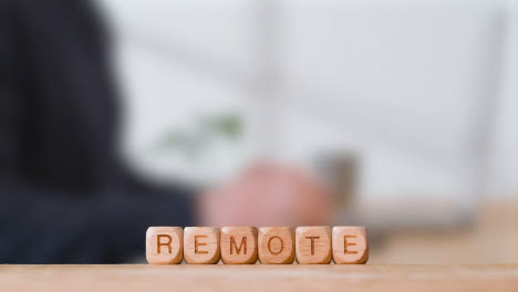 Business-Concept-Wooden-Letter-Cubes-Or-Dice-Spelling-Remote-With-Office-Person-Using-Mobile-Phone-In-Background