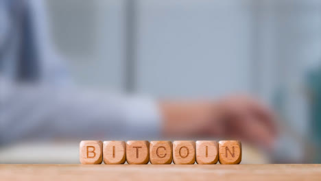 Business-Concept-Wooden-Letter-Cubes-Or-Dice-Spelling-Bitcoin-With-Office-Person-Working-At-Laptop-In-Background