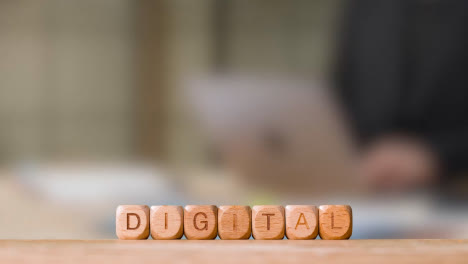 Business-Concept-Wooden-Letter-Cubes-Or-Dice-Spelling-Digital-With-Office-Person-Working-At-Laptop-In-Background