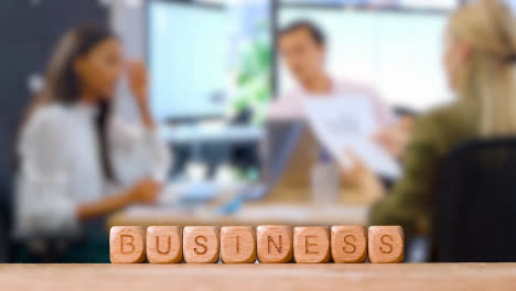 Business-Concept-Wooden-Letter-Cubes-Or-Dice-Spelling-Business-With-Office-Meeting-In-Background