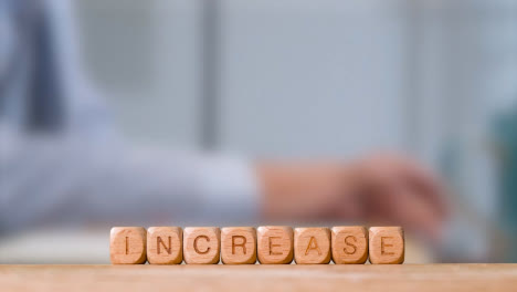 Business-Concept-Wooden-Letter-Cubes-Or-Dice-Spelling-Increase-With-Office-Person-Working-At-Laptop-In-Background