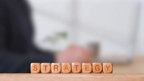 Business-Concept-Wooden-Letter-Cubes-Or-Dice-Spelling-Strategy-With-Office-Person-Using-Mobile-Phone-In-Background