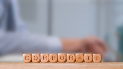 Business-Concept-Wooden-Letter-Cubes-Or-Dice-Spelling-Corporate-With-Office-Person-Working-At-Laptop-In-Background