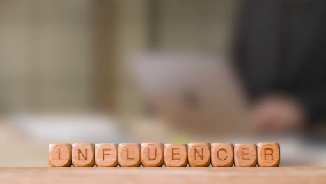 Business-Concept-Wooden-Letter-Cubes-Or-Dice-Spelling-Influencer-With-Office-Person-Working-At-Laptop-In-Background