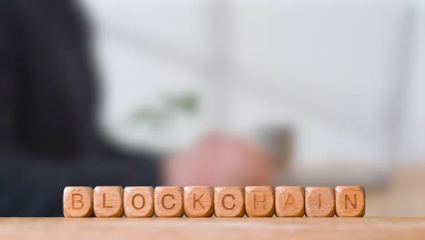 Business-Concept-Wooden-Letter-Cubes-Or-Dice-Spelling-Blockchain-With-Office-Person-Using-Mobile-Phone-In-Background