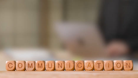 Business-Concept-Wooden-Letter-Cubes-Or-Dice-Spelling-Communication-With-Office-Person-Working-At-Laptop-In-Background
